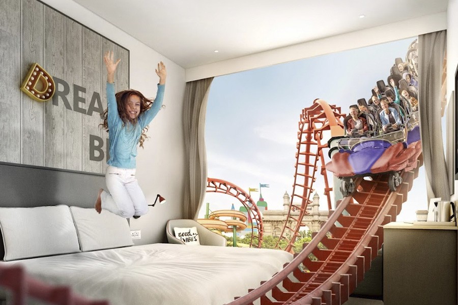 A girl joyfully jumps onto a bed, with a colorful roller coaster in the background at Rove At The Park Hotel, Dubai