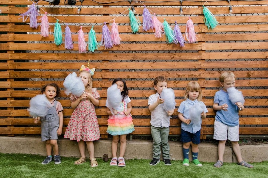 A group of children at a birthday party, smiling in front of a wooden fence, each holding colorful cotton candy