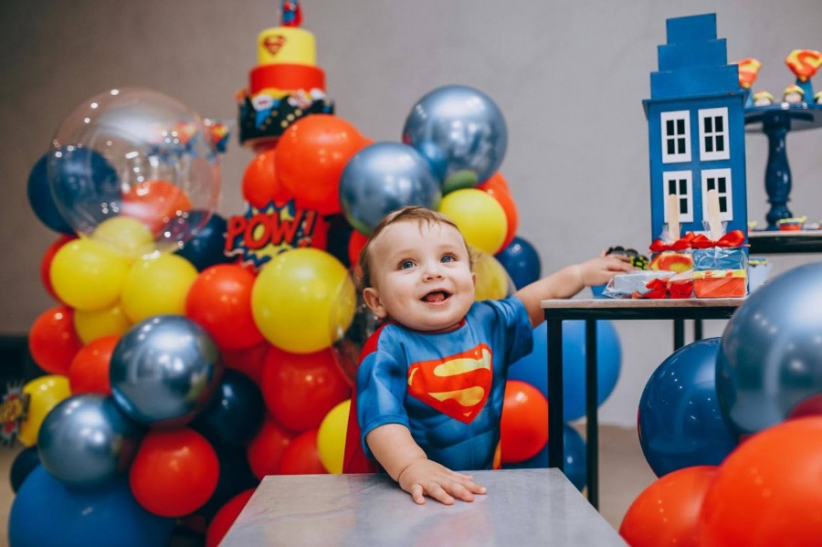 A baby in a Superman costume sits on a table surrounded by colorful balloons at a birthday party