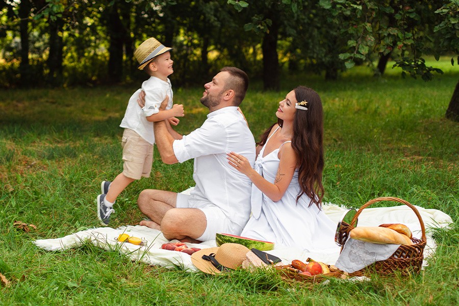 A family enjoying a picnic on a blanket in a sunny park, surrounded by trees and playful children