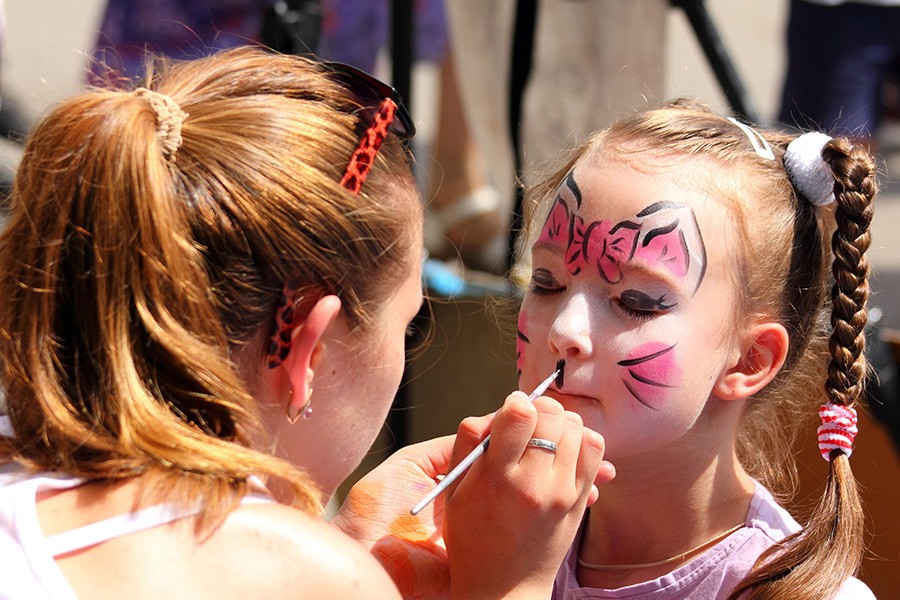 A girl smiles as a face painter applies a cat design on her cheek at a family-friendly event